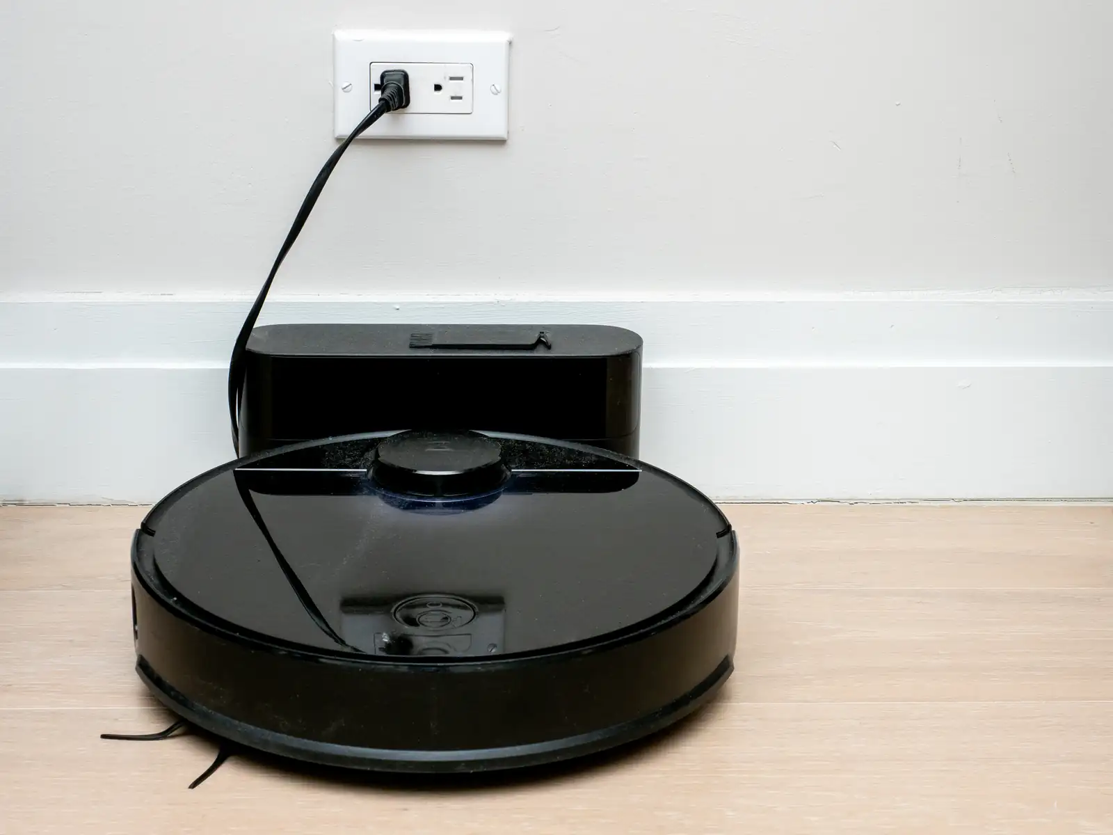 Foto: ajay_suresh · CC BY 2.0 · Wikimedia Commons Robot aspirador negro apoyado en su base de carga, pegada al rodapié y enchufada a la pared